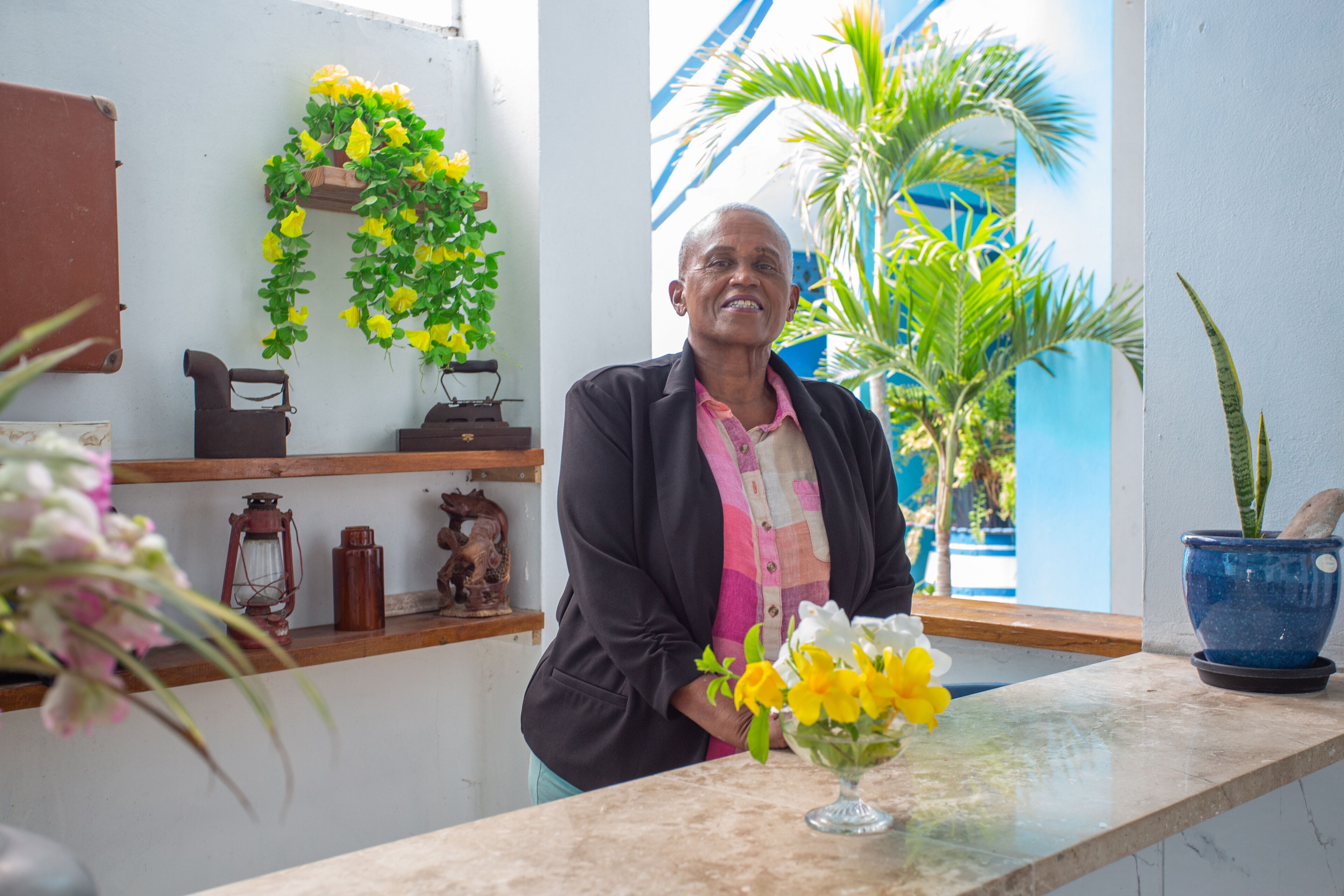 Un hombre de pie frente a un mostrador con un florero, estantes con objetos decorativos y una pared azul con una planta.