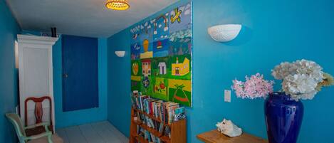 A room with a blue wall, a bookshelf filled with books, a wooden chair, a blue vase with flowers, and a colorful mural.