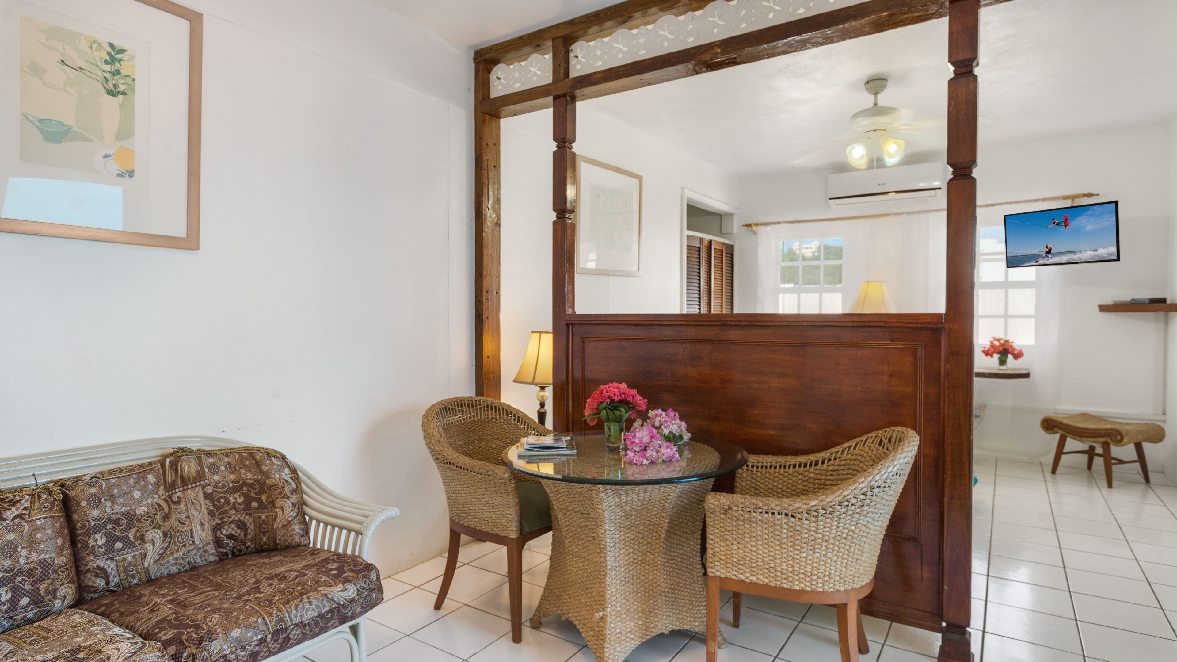 A room with a sofa, wicker chairs, a glass table with flowers, a wooden partition, a TV, and a ceiling fan.