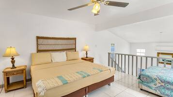 A bedroom with a large bed, two bedside tables with lamps, a ceiling fan, a balcony with a railing, and a view of the sea through a window.