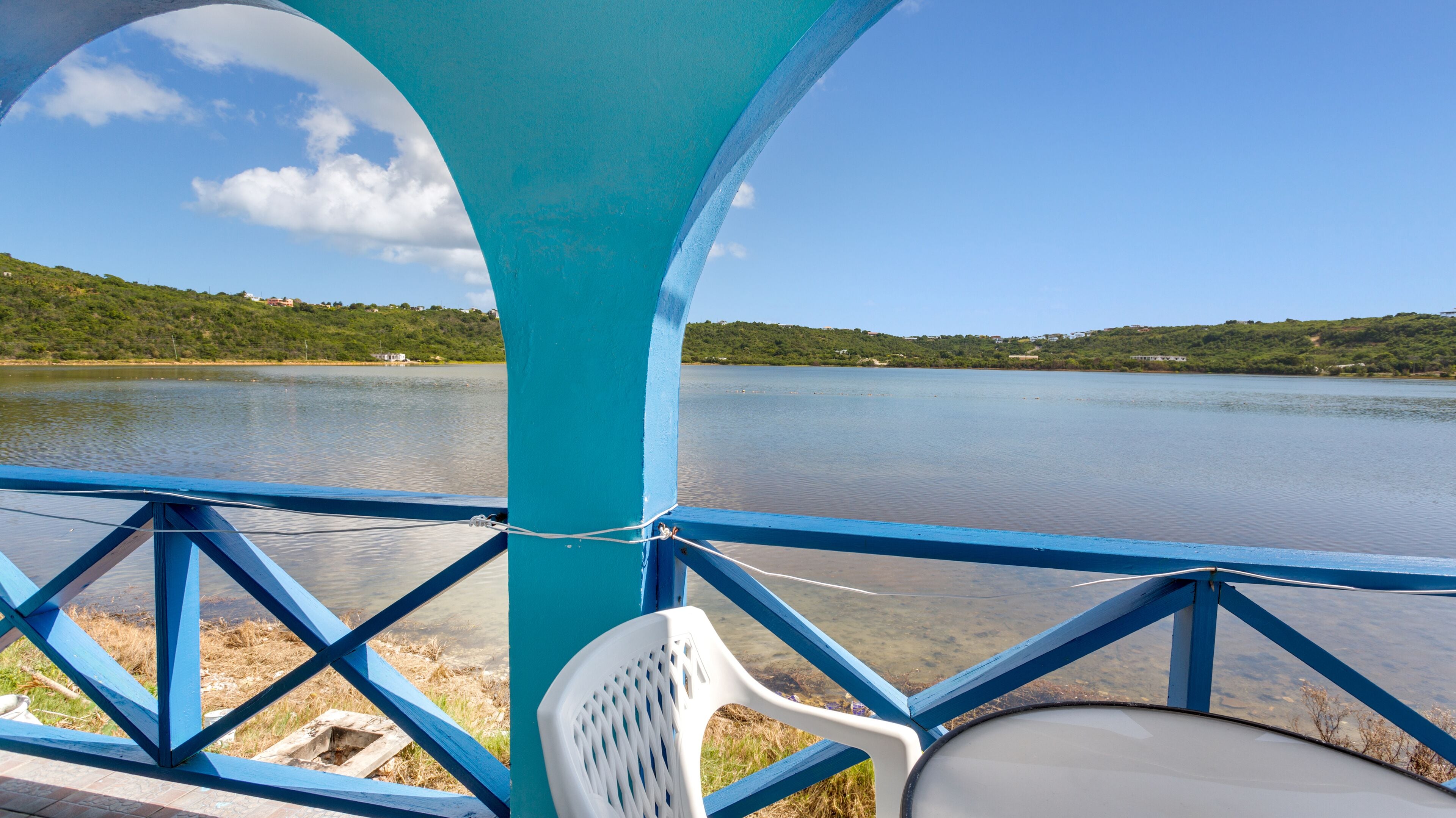 Vista desde una terraza azul que da a un lago tranquilo, con una silla de plástico blanco y una barandilla de metal.