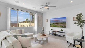 A modern living room with a large window, a ceiling fan, a flat-screen TV, a white sofa, a coffee table, and a side table with a plant. - NEW 9 bd home w/Pergola|Large Gameroom|Huge Pool (Coachella)
