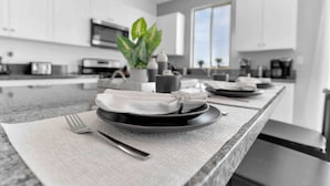 A modern kitchen with a central island, a plant, and a dining setup with plates and cutlery. - NEW 9 bd home w/Pergola|Large Gameroom|Huge Pool (Coachella)