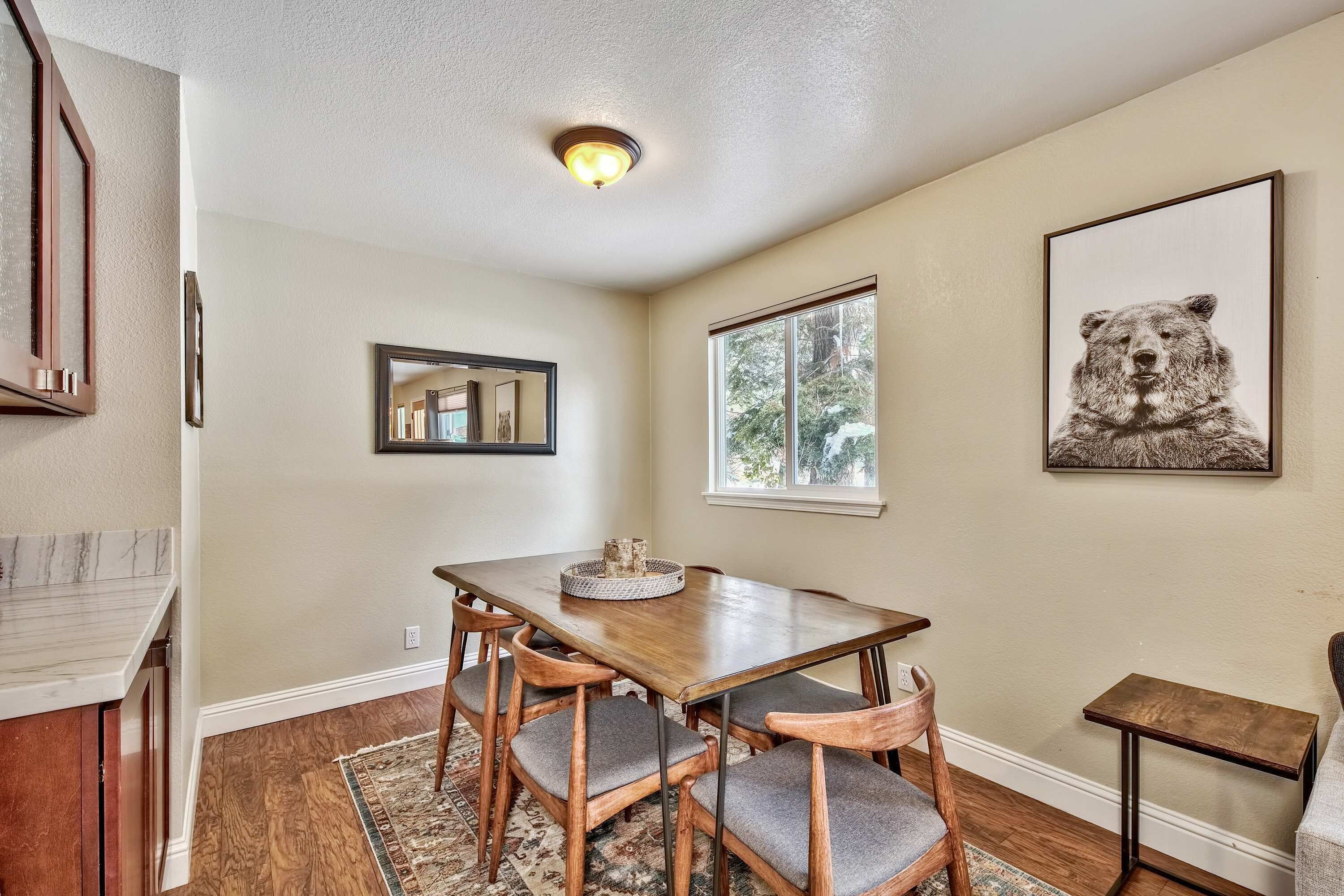 A dining area with a wooden table, four chairs, a rug, a small side table, and a bear artwork on the wall.