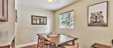 A dining area with a wooden table, four chairs, a rug, a small side table, and a bear artwork on the wall.
