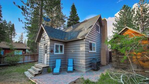 A small, single-story house with a sloped roof, a brick chimney, and a small porch with two blue lawn chairs. - Dancing Bear Cabin Pet Friendly, Sleeps 4, Totally Cute! (South Lake Tahoe)