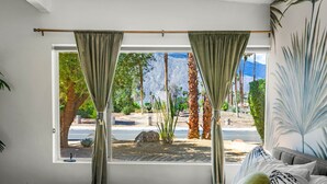 A bedroom with a large window, curtains, a bed with striped bedding, and a view of a landscaped area with trees and a road. - Mid-Century Alexander Pool Home Featured in Modernism Week (Palm Springs)