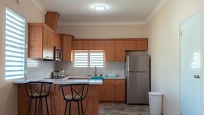 A compact kitchen with wooden cabinets, a stainless steel refrigerator, a microwave, and a sink. There are two bar stools and a window with blinds. - Beachfront Studios (Fajardo)