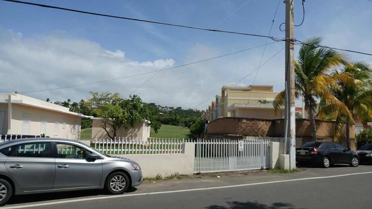 A residential street with a gated entrance, a parked silver car, and a building with a white fence.