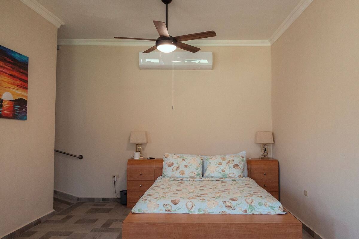 A bedroom with a bed, two bedside lamps, a ceiling fan, and a painting on the wall.