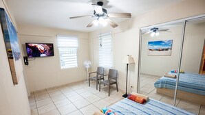 A hotel room with a bed, a chair, a TV, a ceiling fan, and a window with blinds. - Beachfront Studios (Fajardo)