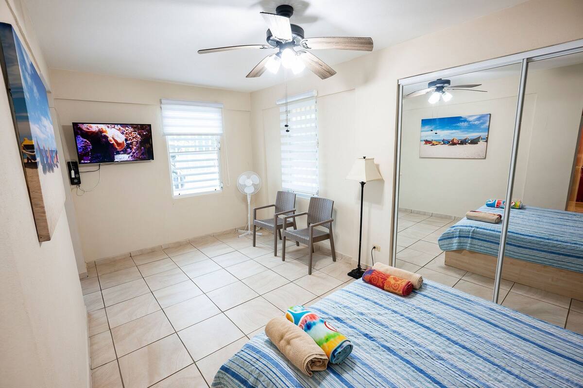 A hotel room with a bed, a chair, a TV, a ceiling fan, and a window with blinds.