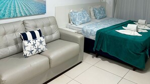 Una habitación de hotel moderna con cama, sofá, un cuadro de playa y baño con cortina de ducha.