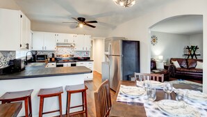 A modern kitchen with white cabinets, a central island, and stainless steel appliances. There is a dining area with a wooden table and bar stools.