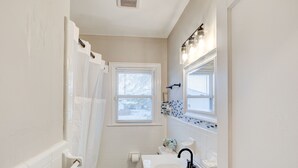 A bathroom with a white bathtub, a sink, and a toilet. There is a window with blinds and a mirror above the sink.