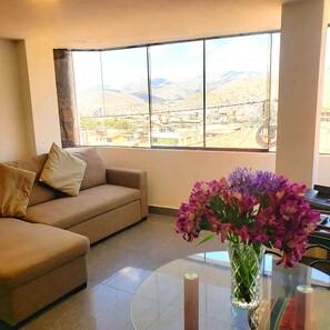 A living room with a beige sofa, a glass coffee table, and a vase with purple flowers. A large window offers a view of a cityscape and mountains. - PRIVATE APARTMENT WITH AMAZING VIEW OF CUSCO (Cusco)