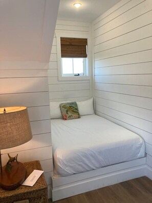 Una habitación pequeña con cama individual, paredes blancas, una ventana con persiana de madera, una lámpara de noche y una mesita con lámpara y un adorno.