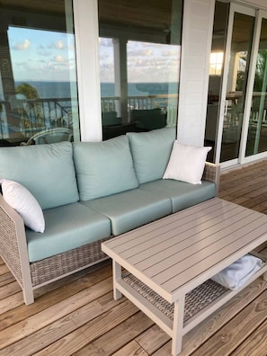 A light-colored wicker sofa with cushions, a matching wooden table, and a glass door leading to an outdoor area with a view of the sea.