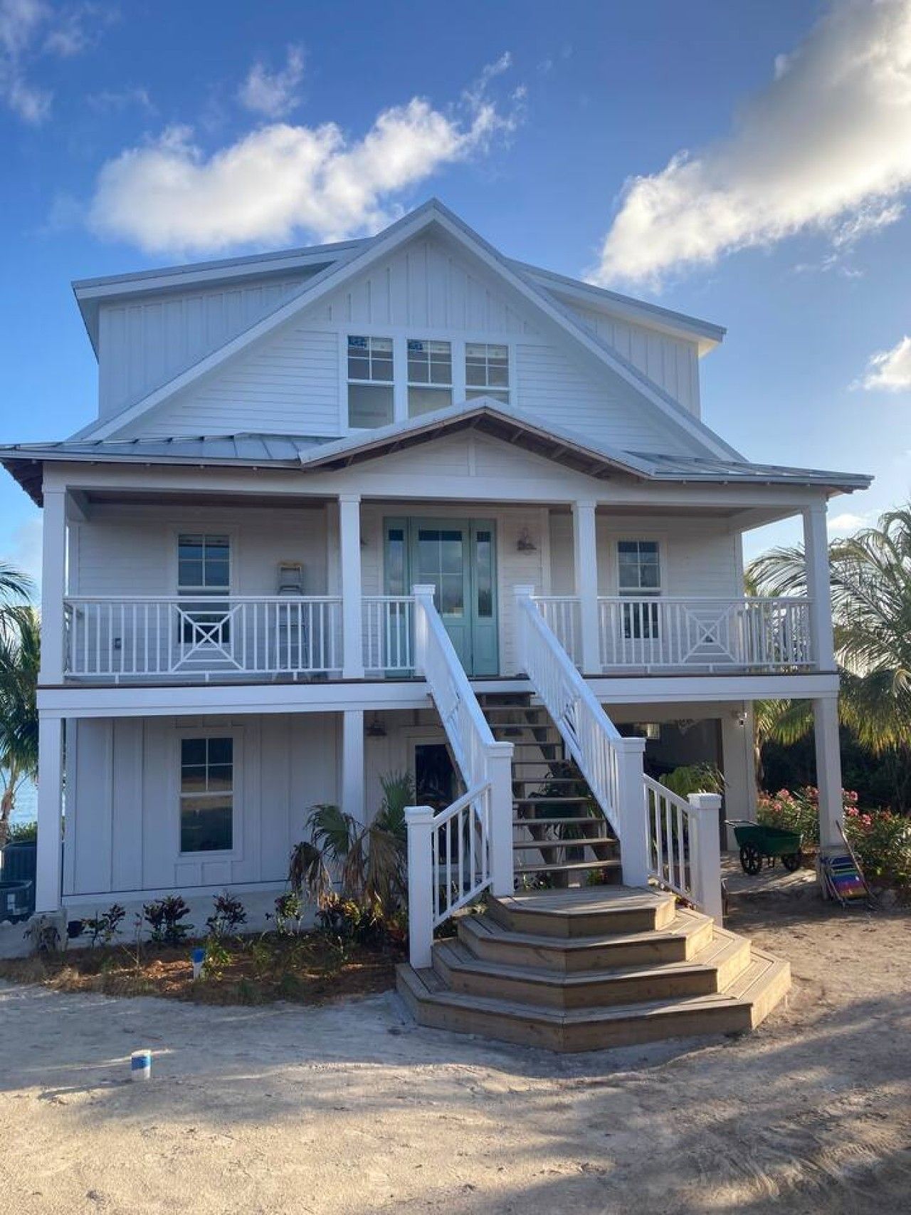 A two-story beachfront house with a balcony, a staircase leading to the entrance, and a sandy ground.