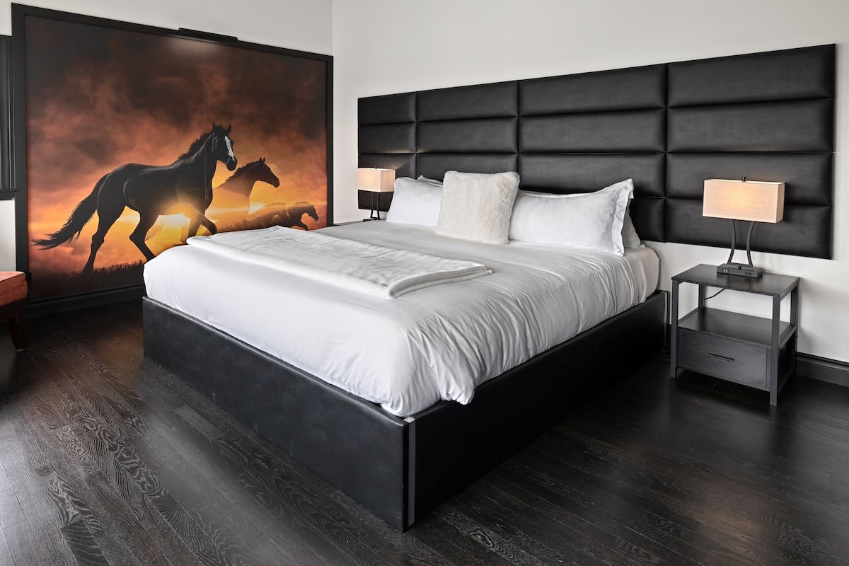 A modern bedroom with a large bed, dark wood flooring, a black headboard, two bedside tables with lamps, and a large painting of horses on the wall.