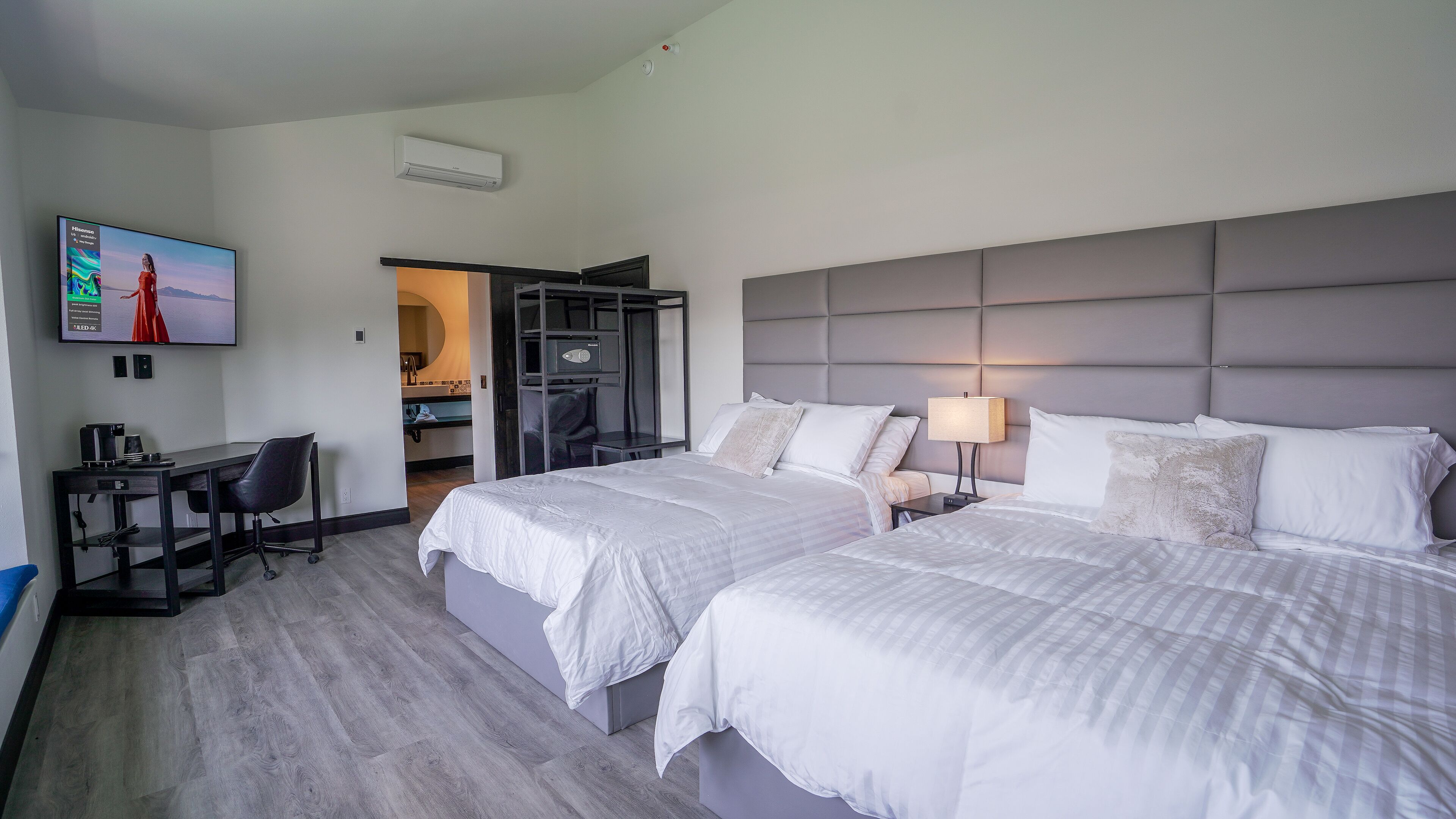 A hotel room with two beds, a desk, a chair, a TV, and a bathroom visible through an open door.