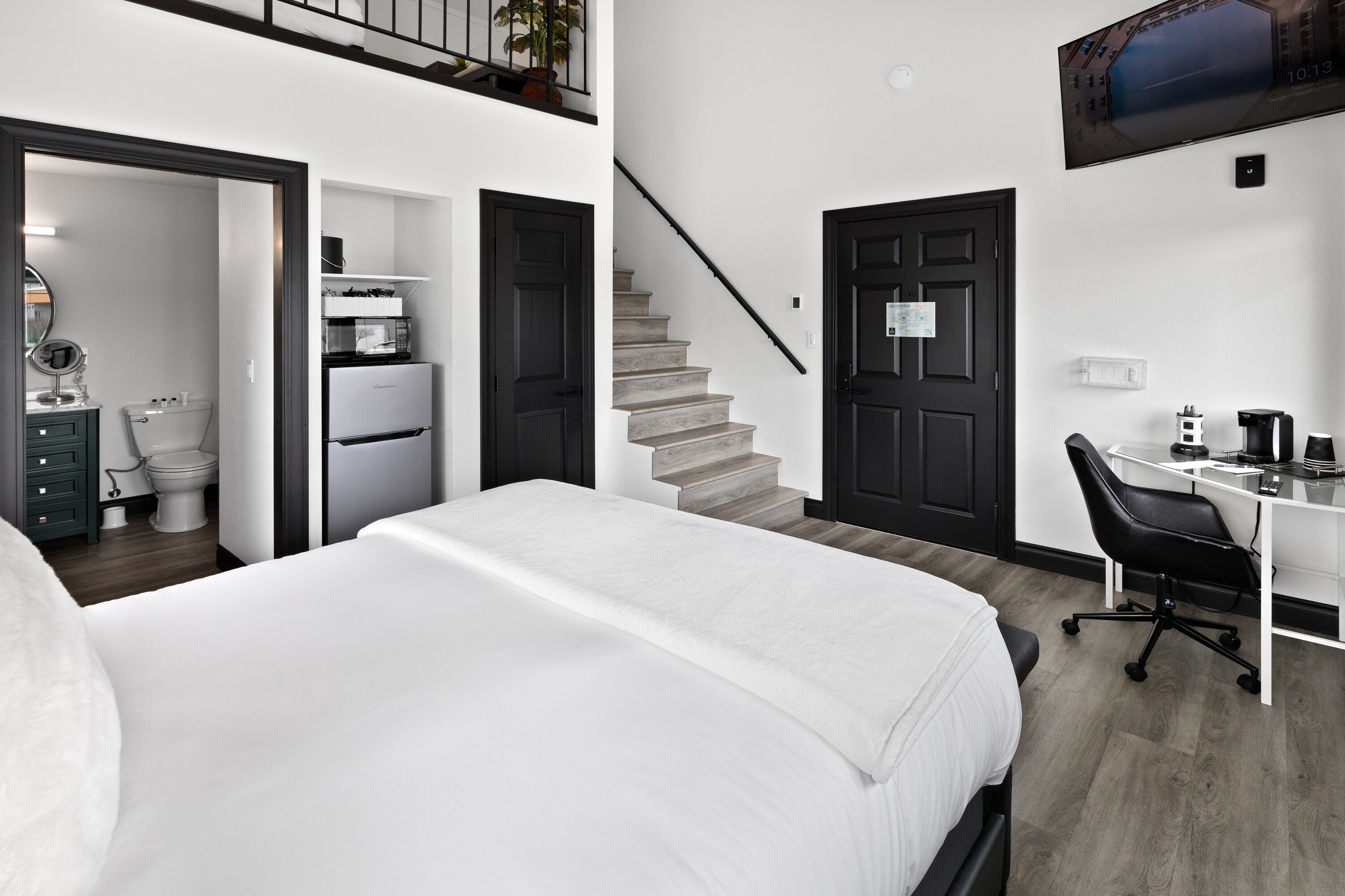 A modern bedroom with a large bed, a desk with a chair, a staircase, and a bathroom visible through an open door.