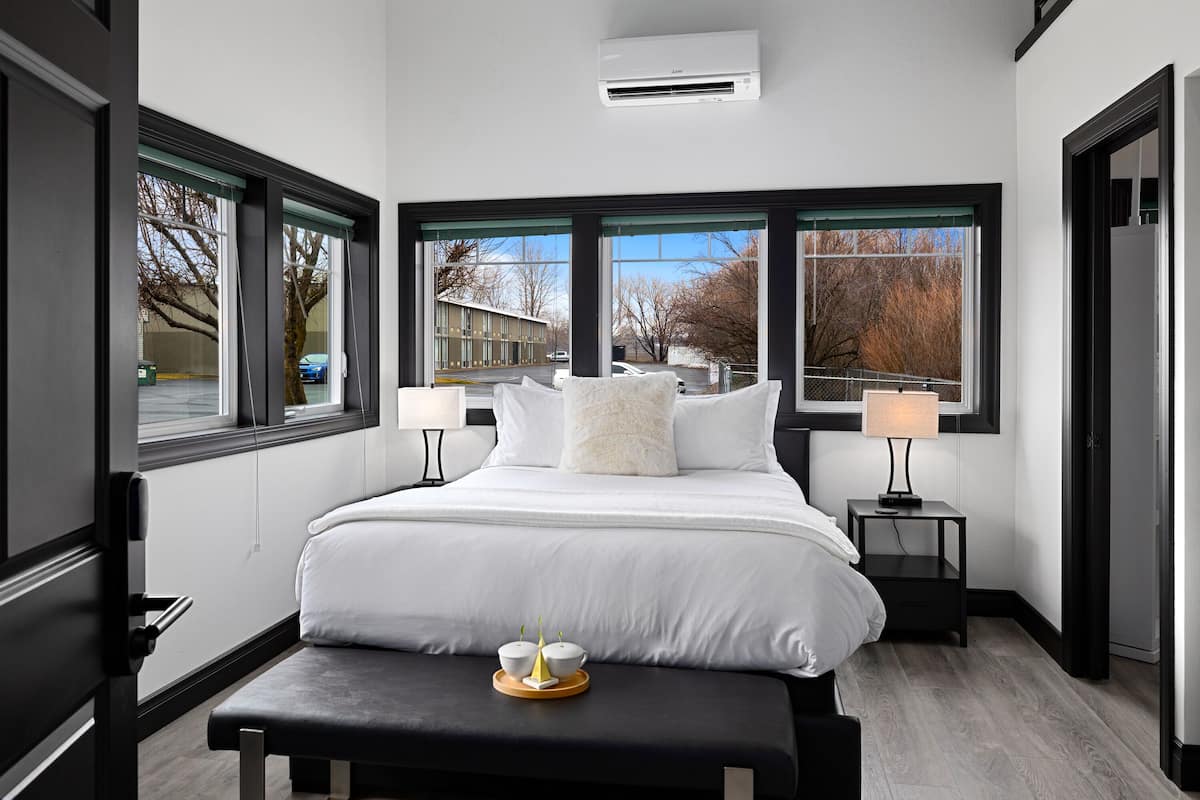 A modern hotel room with a large bed, two bedside tables, and a view of the outside through large windows.
