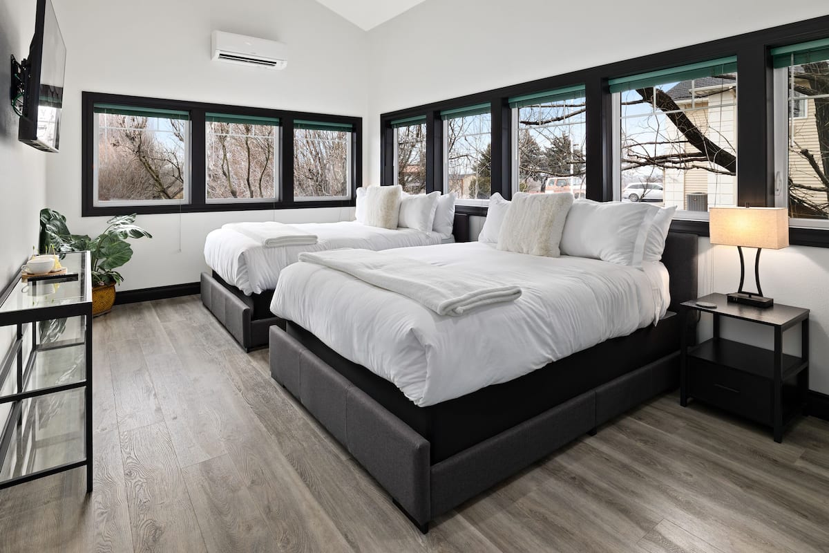 A modern bedroom with two beds, large windows, a bedside table, and a plant.