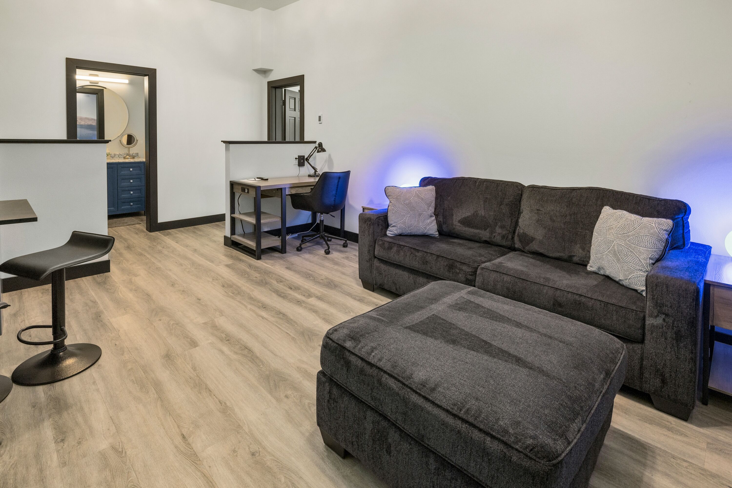 A modern living room with a grey sectional sofa, a small desk with a chair, and a bar stool.