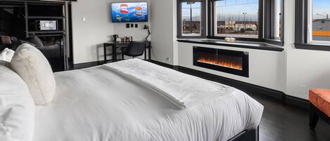 A modern hotel room with a large bed, a fireplace, a TV, and a view of the city through large windows.