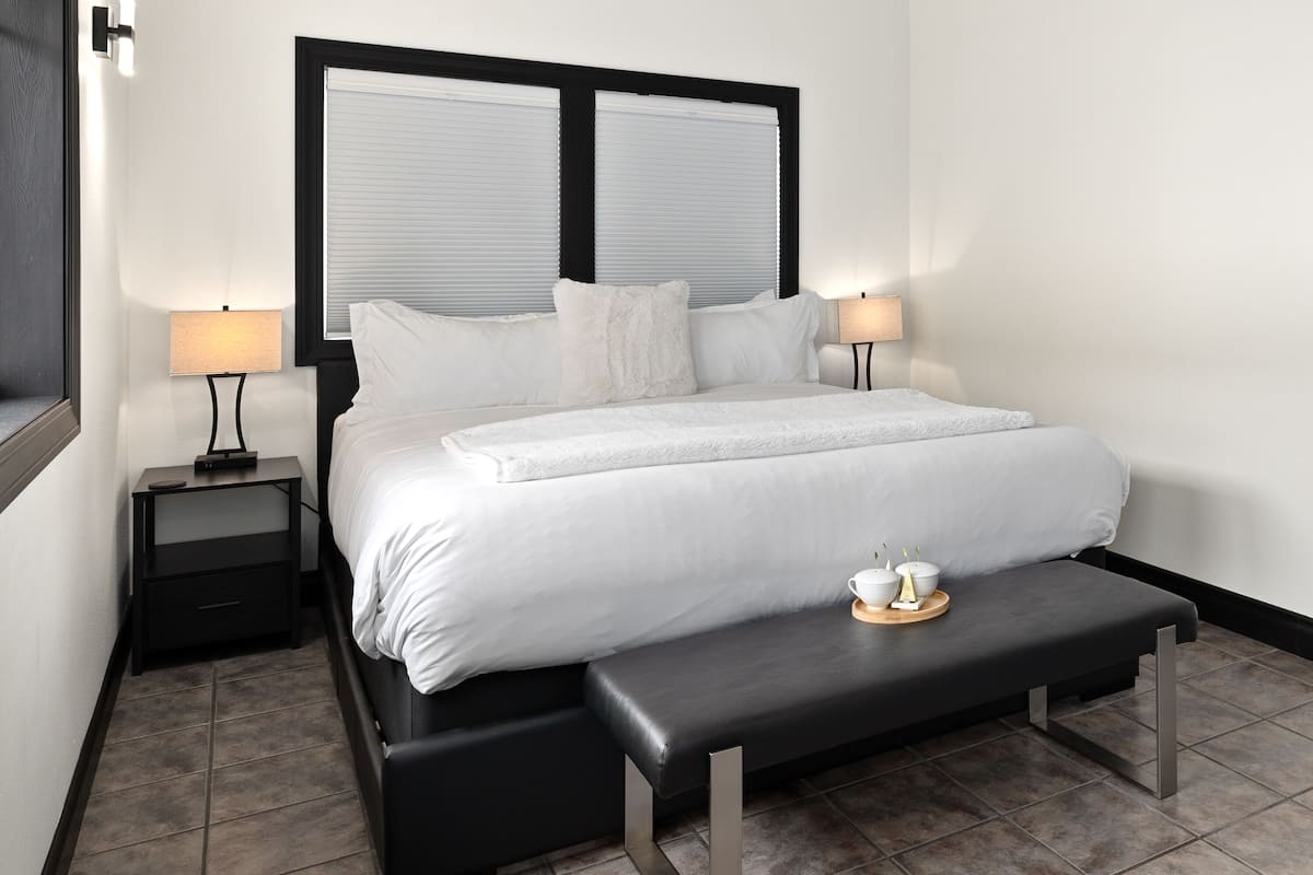 A modern hotel room with a large bed, a bedside table with a lamp, a bench with a tray of items, and a window with blinds.