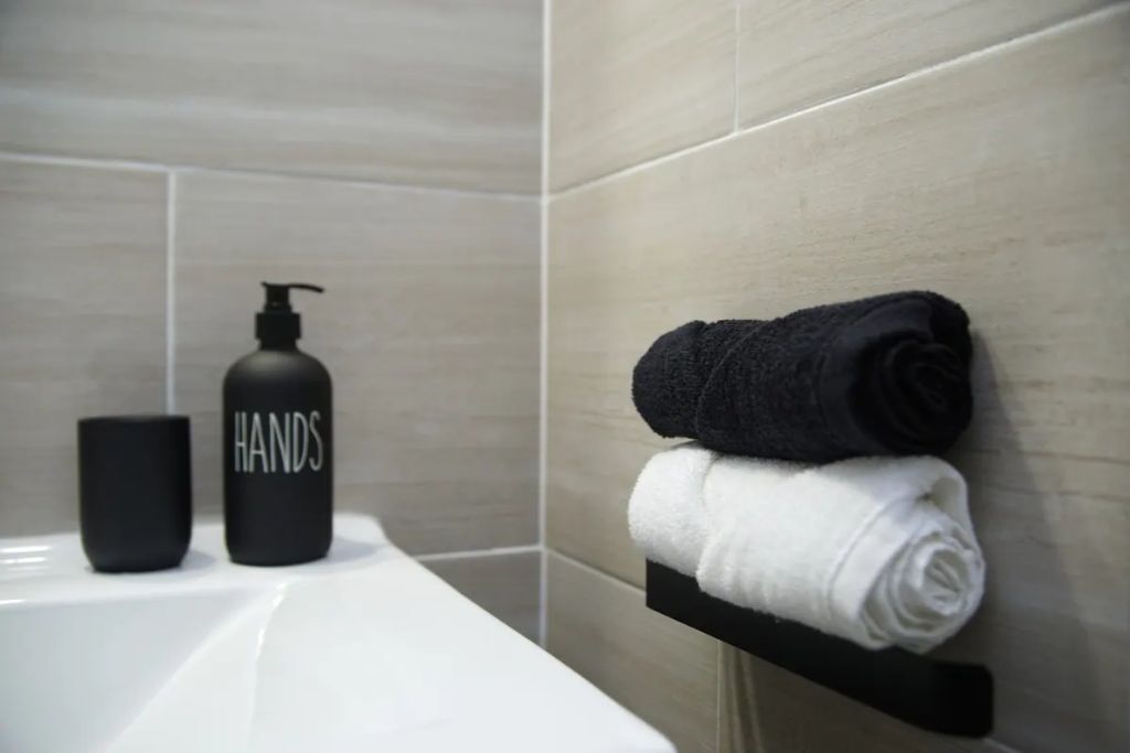 Une salle de bain avec un lavabo blanc, un distributeur de savon noir portant l’inscription « MAINS », et deux serviettes, une noire et une blanche, suspendues à un porte-serviettes noir.