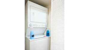 Lavadora blanca compacta con dos botellas azules de detergente, ubicada en un lavadero pequeño con una puerta y persianas blancas.