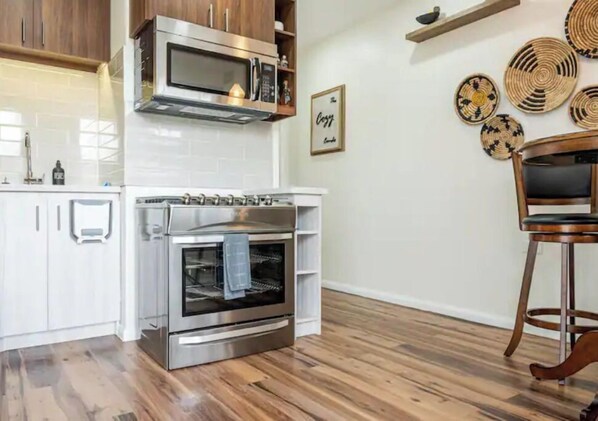 A kitchen with white cabinets, a stainless steel oven, and a microwave. There are woven baskets on the wall and a wooden chair. - "Modern Comfort Near Airport & Shopping - The Cozy Condo" (Arouca, Tunapuna/Piarco Regional Corporation)