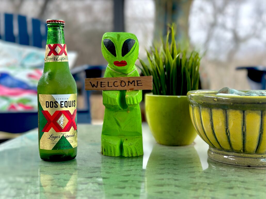 Een fles Dos Equis bier, een groene buitenaards figuurtje, een welkomstbord, een potplant en een gele schaal op een tafel.