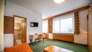 A room with a green patterned floor, a wooden table, an orange sofa, a dining table with chairs, a television on the wall, and a window with curtains.