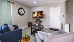 A living room with a sofa, a coffee table, a TV displaying a fireplace, a clock, and a door.