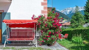 Un banco rojo con toldo blanco, un arbusto de flores rosas y montañas al fondo.