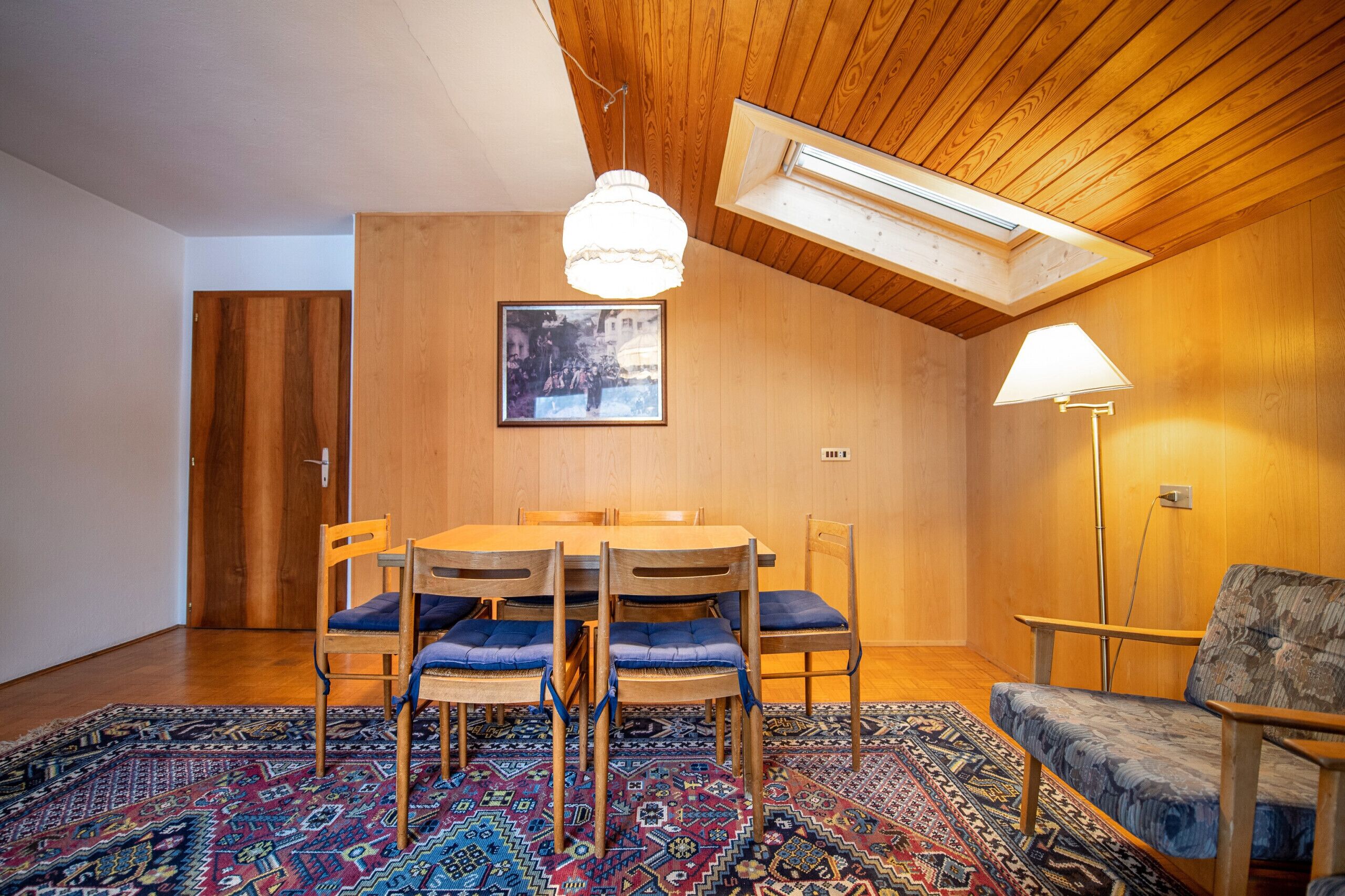 En matplats med ett träbord och stolar, en mönstrad matta, en taklampa och en vägglampa.