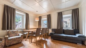 Ein großzügiger Wohnbereich mit Sofa, Sesseln, einem Esstisch und großen, mit Vorhängen versehenen Fenstern.