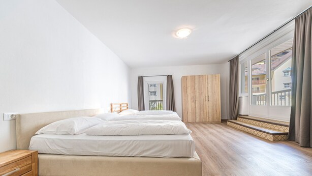 Ein modernes Schlafzimmer mit einem großen Bett, Holzboden, einem Nachttisch, einem Kleiderschrank und Blick auf ein Gebäude durch ein großes Fenster.