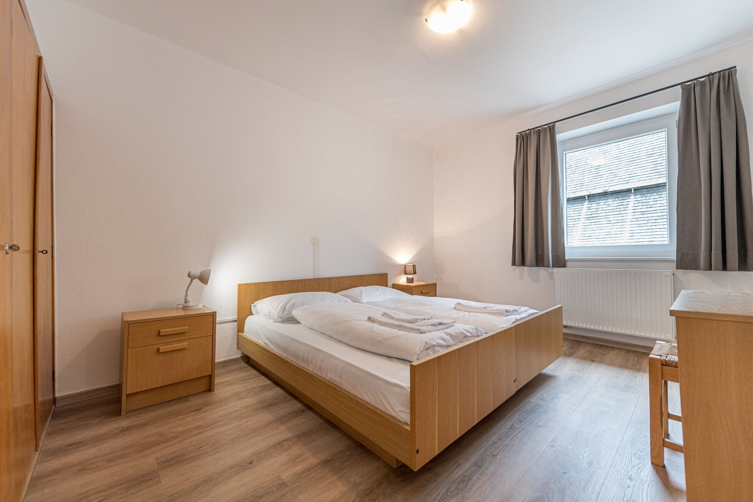 Une chambre à coucher avec un lit en bois, des tables de chevet, une commode, une fenêtre avec des rideaux et un radiateur.
