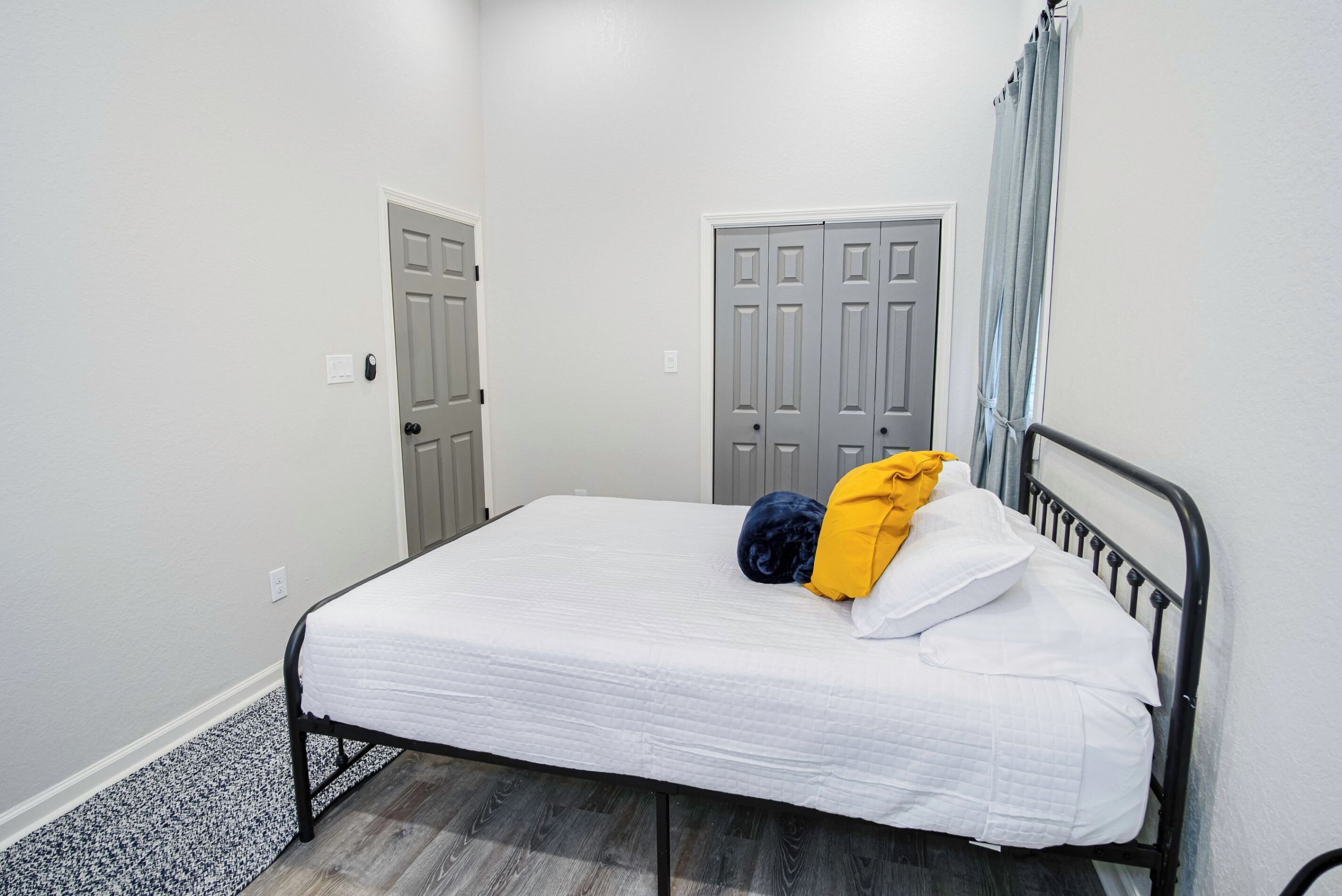 Un lit simple avec un matelas blanc et un cadre en métal noir, une porte grise et une armoire grise avec un rideau.