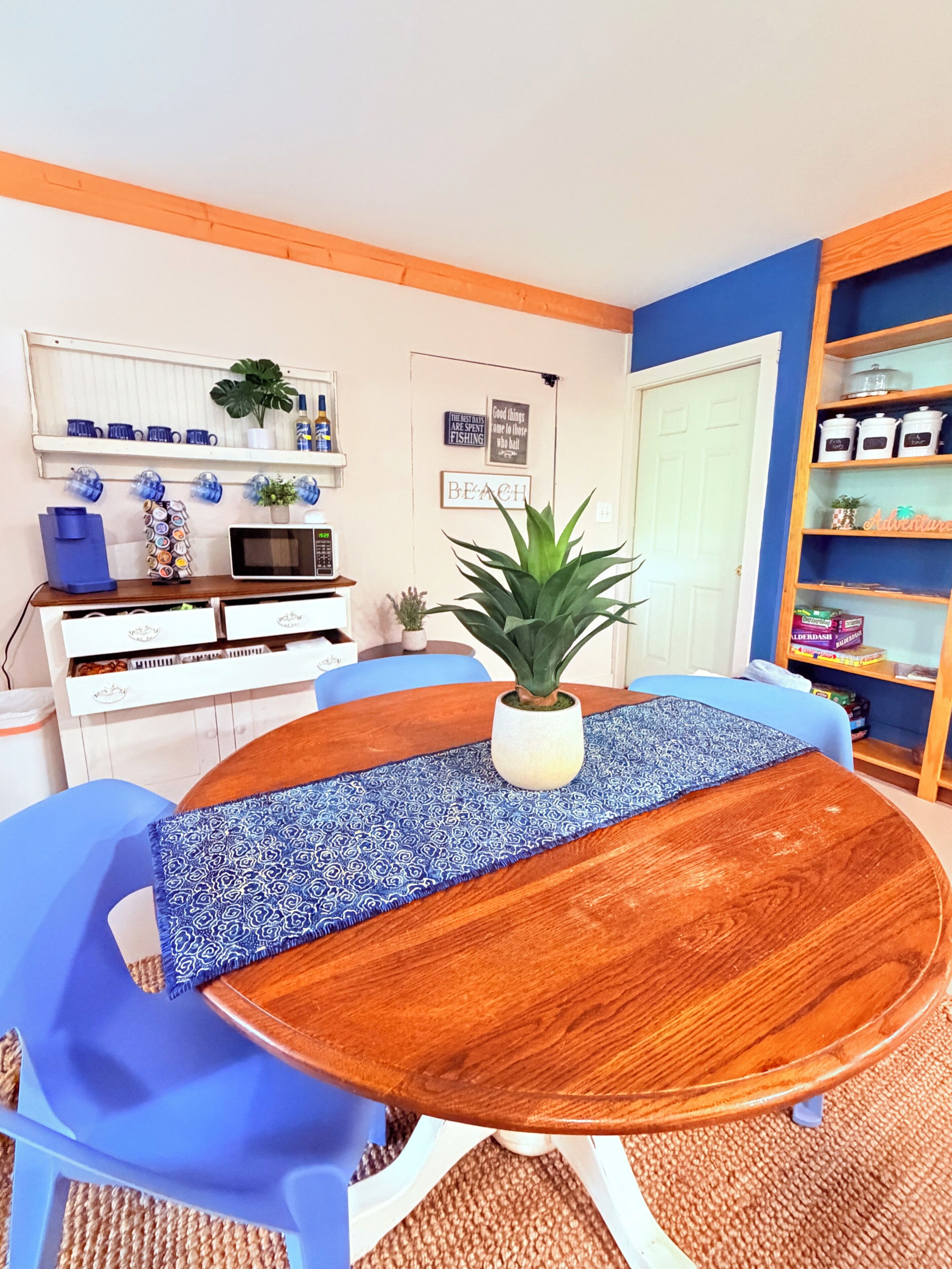 A dining area with a wooden table, blue chairs, a potted plant, and a kitchen area with shelves and appliances.