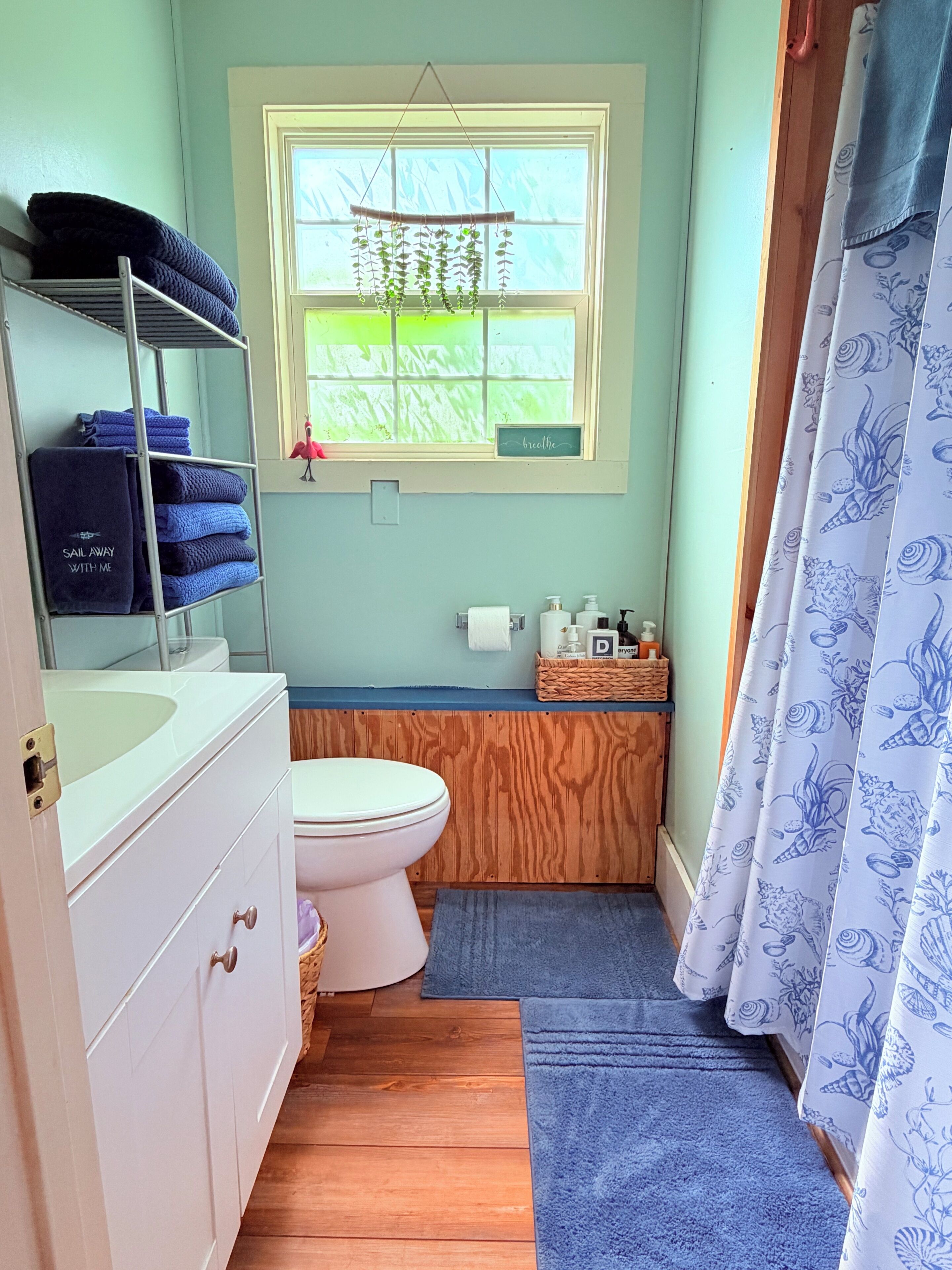A bathroom with a wooden vanity, a white toilet, a window with a view, and a blue shower curtain.