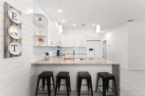 A modern kitchen with white cabinetry, a granite countertop, and three black bar stools. - Busch Garden New House (Tampa)