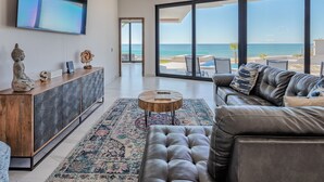 A modern living room with a leather sofa, a wooden console, and a large TV. A view of the ocean is visible through the large windows. - Luxury Oceanfront Home with Pool & Hot Tub - located on Sandy Beach (Puerto Peñasco)