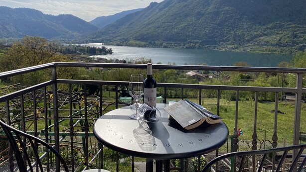 A balcony with a table, a wine bottle, and a book, overlooking a lake and mountains. - 2 vacation apartments for up to 6 people, perfect for a vacation with family or friends (Idro)