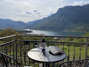Un balcón con una mesa, una botella de vino y un libro, con vistas a un lago y montañas.