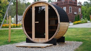 A large wooden barrel structure with a door, set on a gravel area with a house in the background. - Family Villa Sauna and Trails White Mountains (Bethlehem)